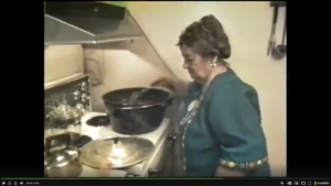 RENE CAISSE IN A GREEEN DRESS STANDING BY HER KITCHEN STOVE AND STIRRING A GRANITE KETTLE OF ESSIAC TEA