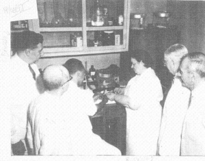 RENE CAISSE’S IS STANDING AND SHOWING SOMETHING ON A COUNTER TO DR BRUSCH AND FOUR OTHER DOCTORS IN WHITE LAB COATS